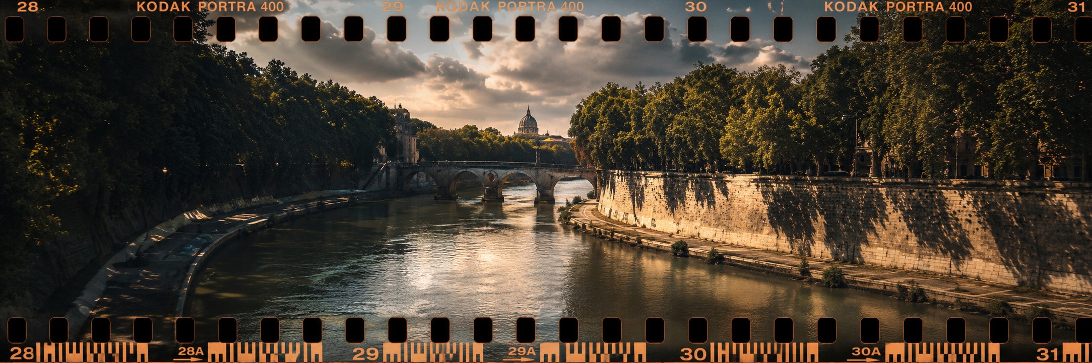 Panoramic film-frame photograph of a calm river bordered by trees, reflecting warm light beneath a cloudy sky.