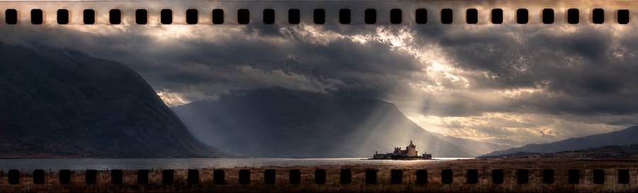 Panoramic film-frame landscape of dark mountains beneath dramatic clouds, with shafts of sunlight reaching the valley below.