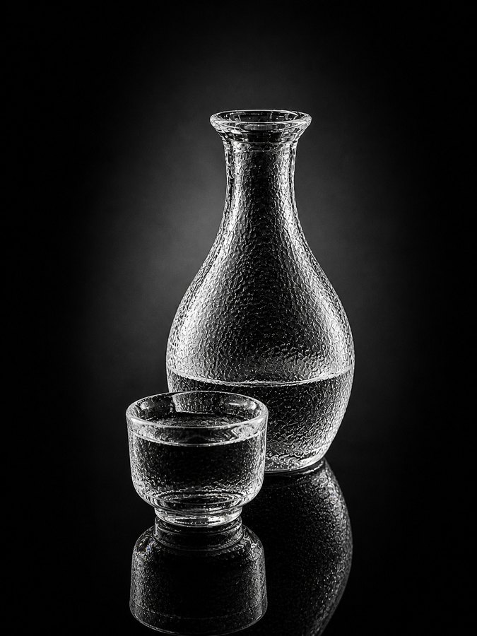 A black-and-white still life shows a textured sake bottle and small cup reflected on a glossy dark surface.
