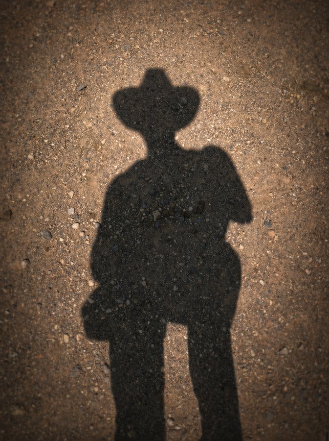 Shadow of a person wearing a broad-brimmed cowboy hat cast across sunlit pavement.