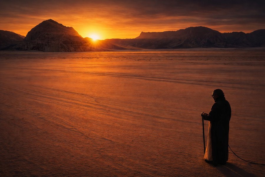 A lone robed figure stands on a vast desert plain at sunset, facing distant mountains beneath an orange sky.