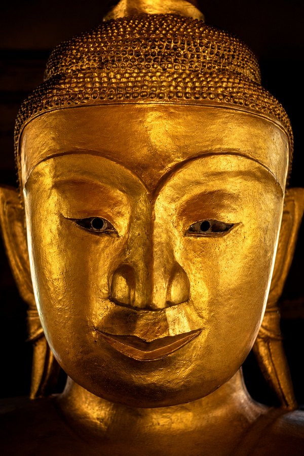 A close-up golden Buddha face fills the frame against a dark background, emphasizing calm expression and metallic texture.