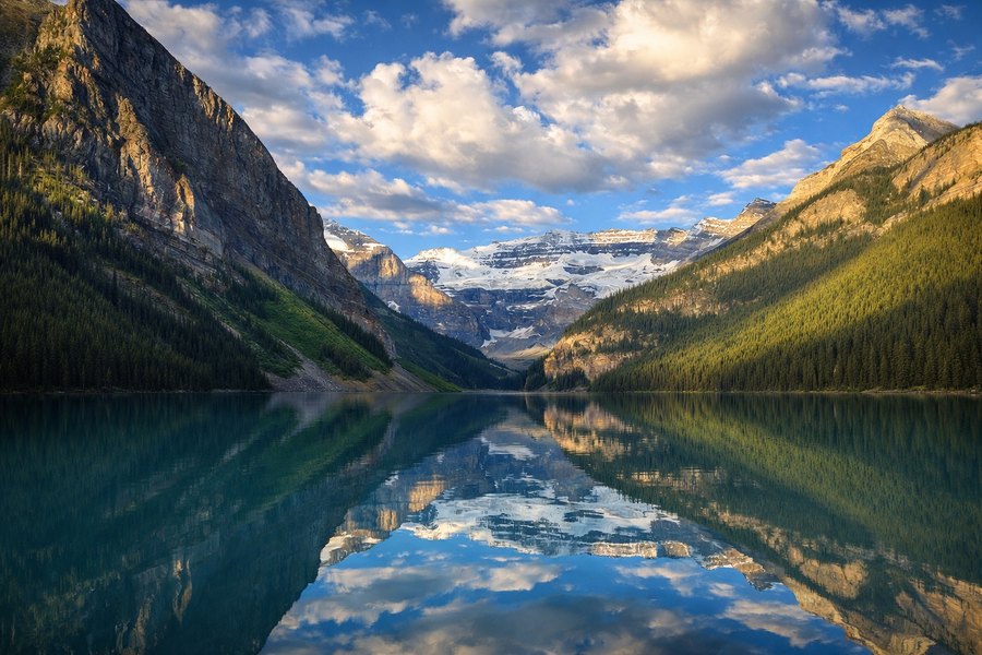Turquoise mountain lake reflecting snowcapped peaks, forested slopes, and a partly cloudy blue sky.