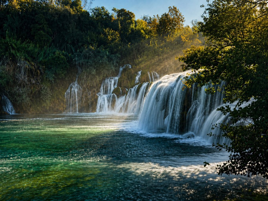 Wide curtain of waterfalls cascading into a clear turquoise pool surrounded by dense green forest.