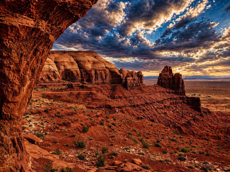 Expansive red-rock desert canyon viewed from beneath a stone overhang under a dramatic cloud-filled sky.