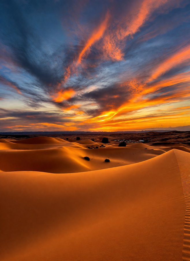 Rolling orange desert dunes beneath streaked blue and red sunset clouds, with small dark shapes in the distance.
