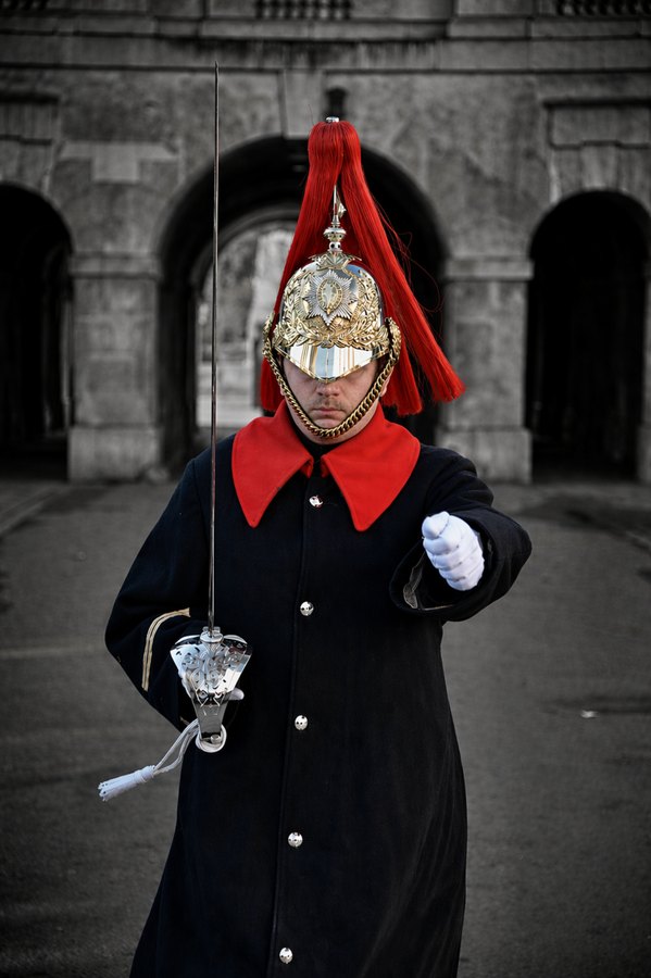 Ceremonial guard in a dark coat and ornate silver helmet with a red plume, holding a sword before stone arches.