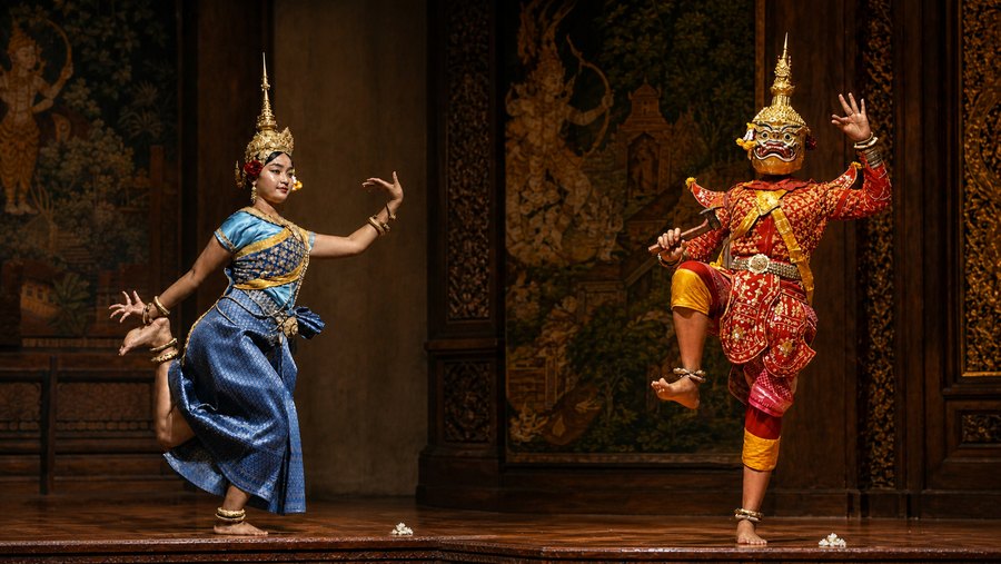 Two traditional Southeast Asian dancers in ornate blue-and-gold and red-and-gold costumes performing on a stage.
