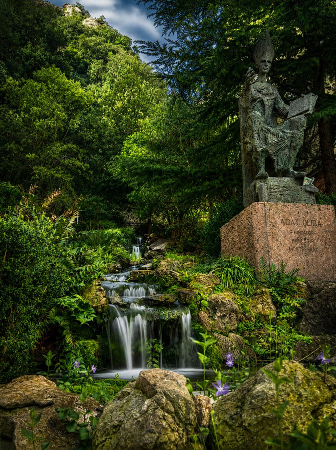 Small garden waterfall flowing through rocks beneath a stone monument, surrounded by lush green trees and plants.