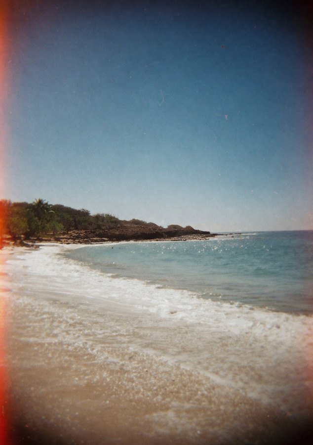Soft-focus vintage photograph of a pale sandy beach and turquoise sea, with a dark tree line and a reddish light leak along one edge.
