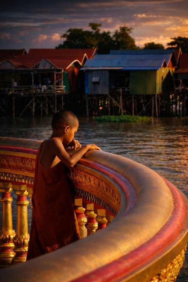 A young Buddhist monk in a saffron robe leans on an ornate curved balcony above a stilt village at sunset.