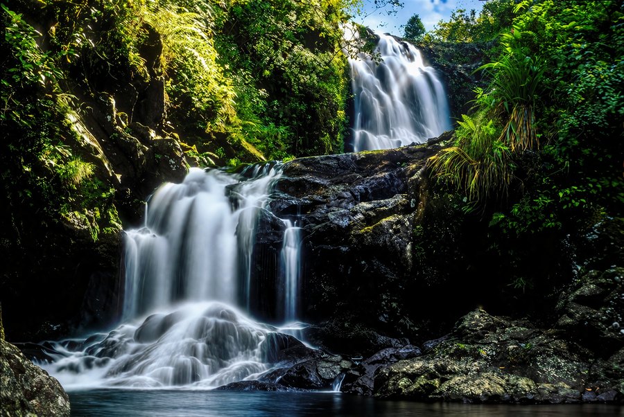 A multi-tiered waterfall pours through dense green forest into a dark pool, photographed with silky long-exposure water.